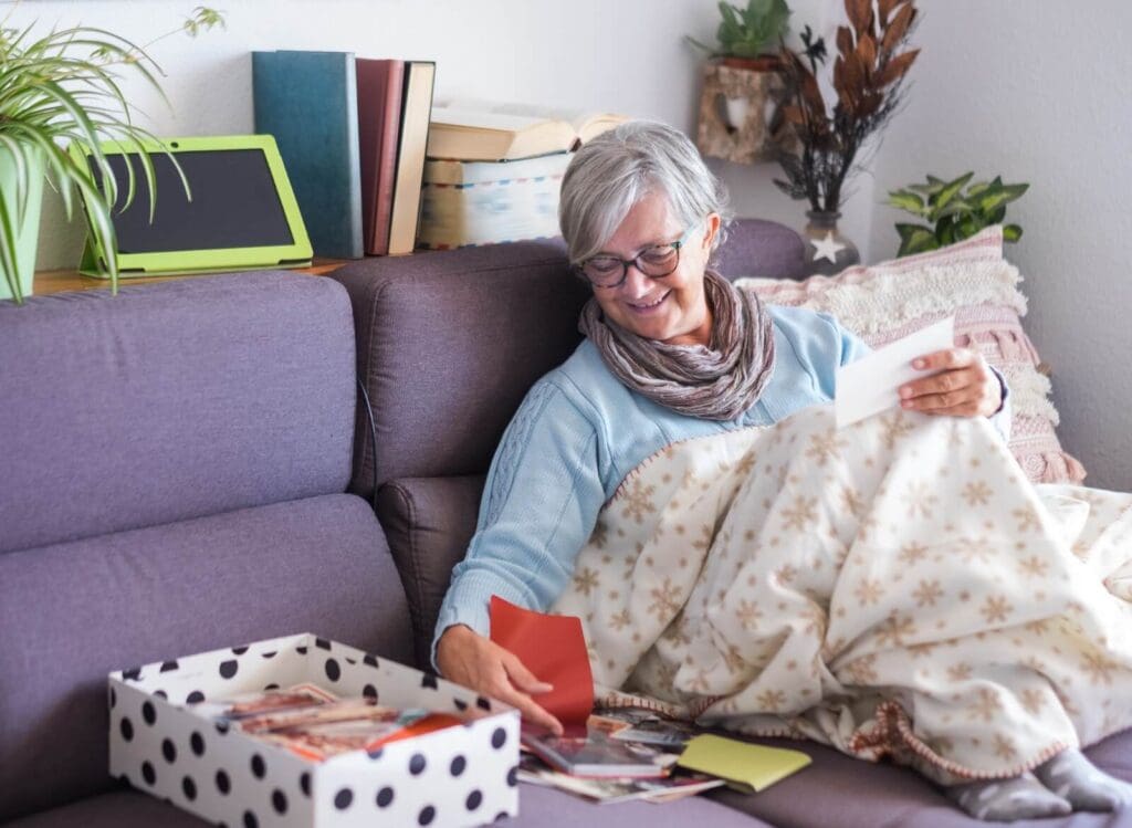 An older adult woman sits on a couch with a blanket over her legs. She is looking at photographs and other items in a small box next to her on the couch. There are plants, books, and a tablet behind her.