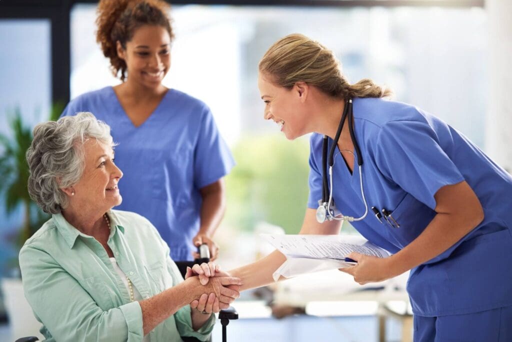 A female health care worker shakes the hand of an older adult woman who is seated in a wheelchair. There is also a female caregiver behind the older woman, pushing the wheelchair.