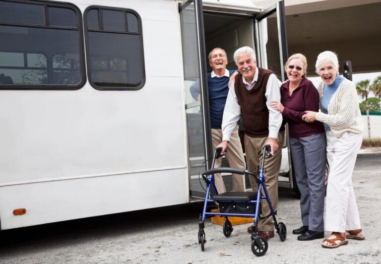 Two older adult men and two older adult women stand outside of a shuttle bus. One man is using a walker.