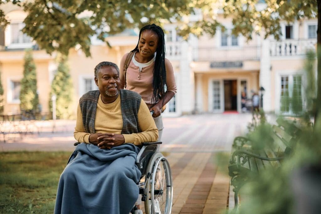 A woman pushes her older adult father in a wheelchair on an outdoor walkway outside a nursing home.
