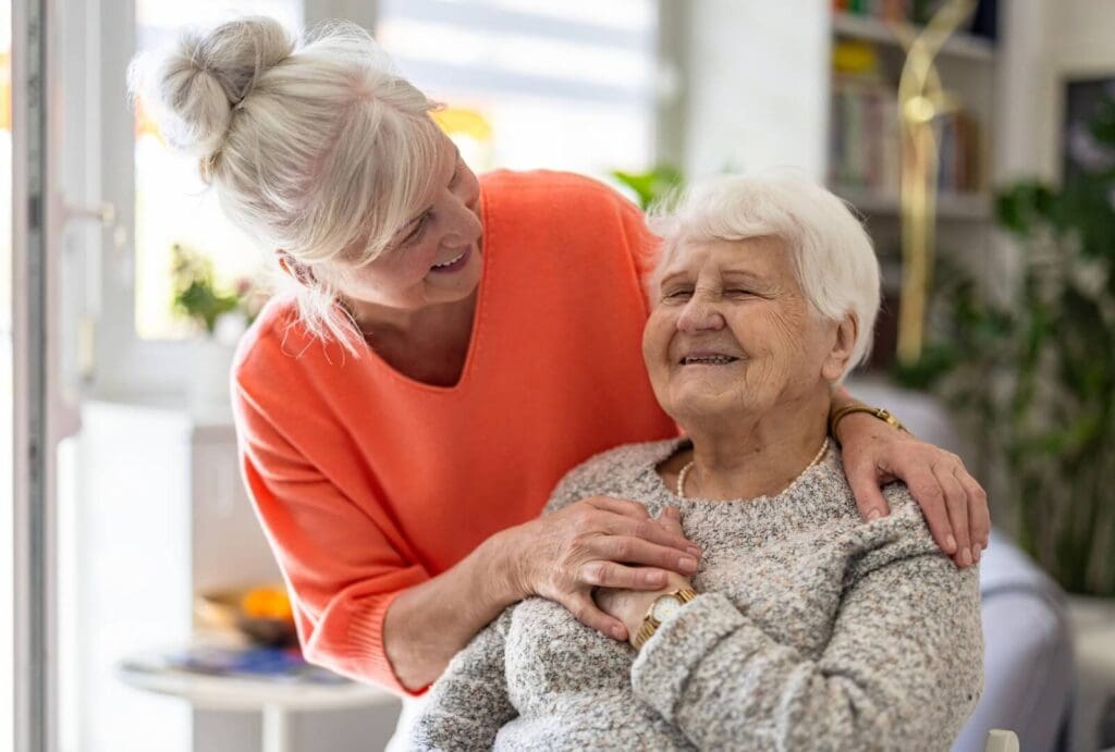 A woman stands behind her seated older adult mother with her hands on her shoulders, leaning down to smile at her.