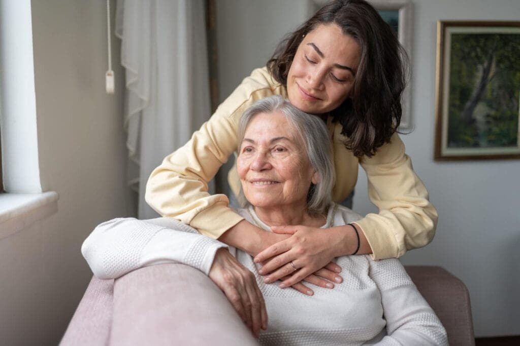 An older adult woman sits on a couch. Her Daughter stands behind her, hugging her. They are smiling.