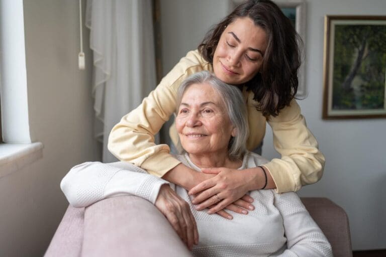 An older adult woman sits on a couch. Her Daughter stands behind her, hugging her. They are smiling.