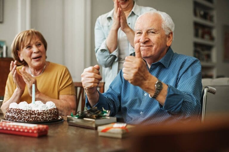 An older adult man is sitting in a wheelchair at a table with a birthday cake on it. There is a woman sitting to his right and a person standing behind him. They are smiling and clapping, and the man is giving a thumbs-up.