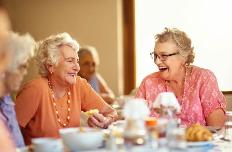 Three older adult women are sitting at a table, enjoying some food together and laughing. There is another older adult woman in the background.