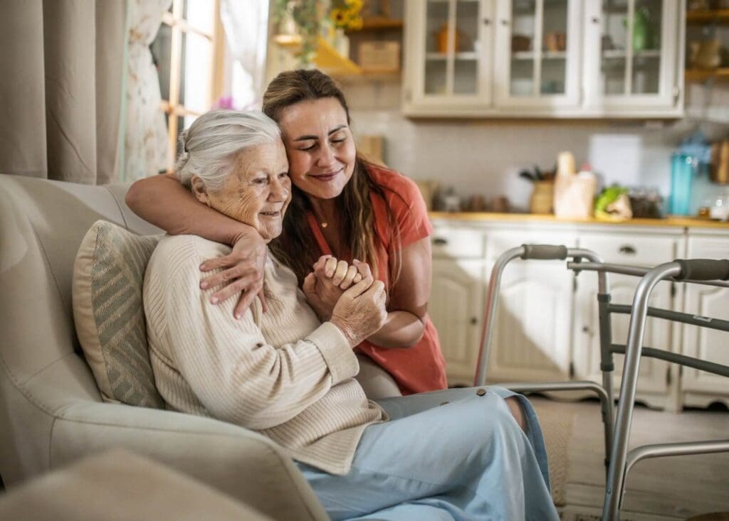 An older adult woman is sitting in an armchair with a walker in front of her. Her daughter is crouching down to hug her. They are smiling.