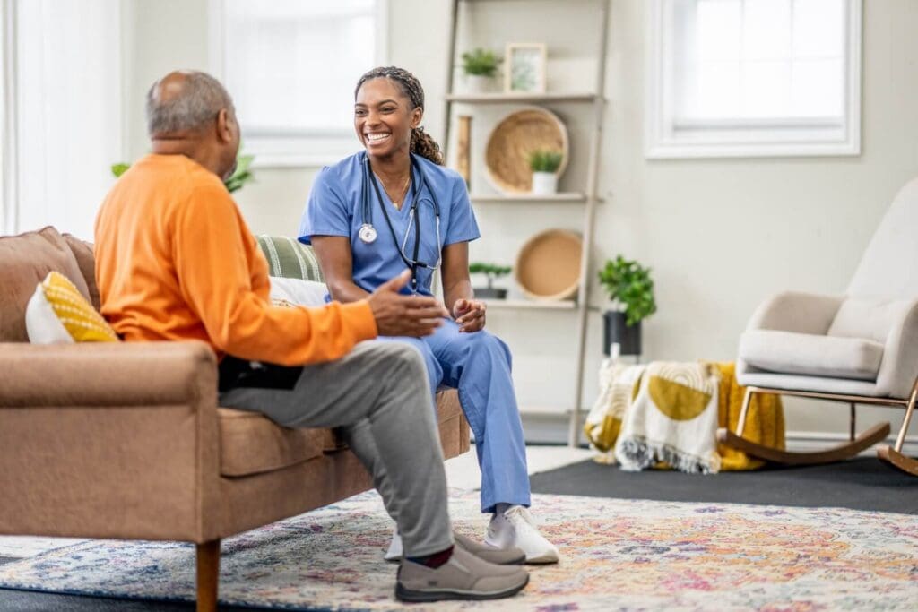 A woman health care working wearing scrubs and a stethoscope sits on a couch with an older adult man who is shown from behind. They are facing each other, and the woman is smiling.