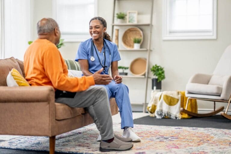 A woman health care working wearing scrubs and a stethoscope sits on a couch with an older adult man who is shown from behind. They are facing each other, and the woman is smiling.