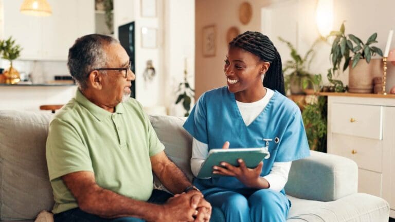 An older adult man sits on a couch with a woman health care worker who is wearing scrubs and holding a tablet.