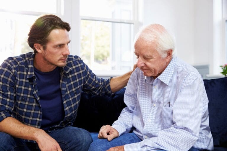 A man sits on a couch with his older adult father with his hand on his shoulder. They look serious.