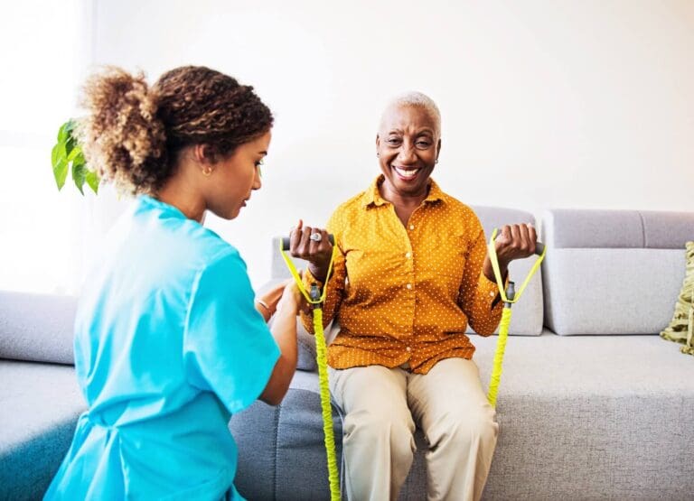 A woman physical therapist is helping an older adult woman who is seated on a couch and holding resistance equipment to rebuild her strength.