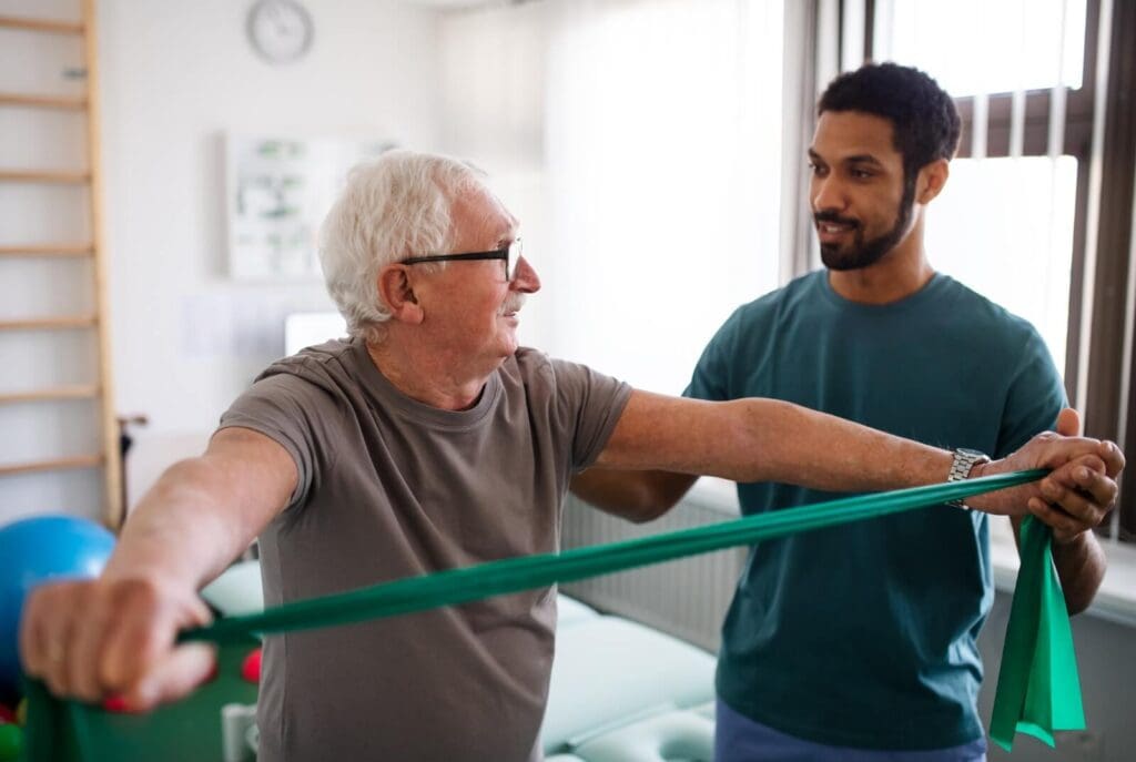 A male physical therapist works with an older adult man. The older man is stretching a resistance band in his hands.
