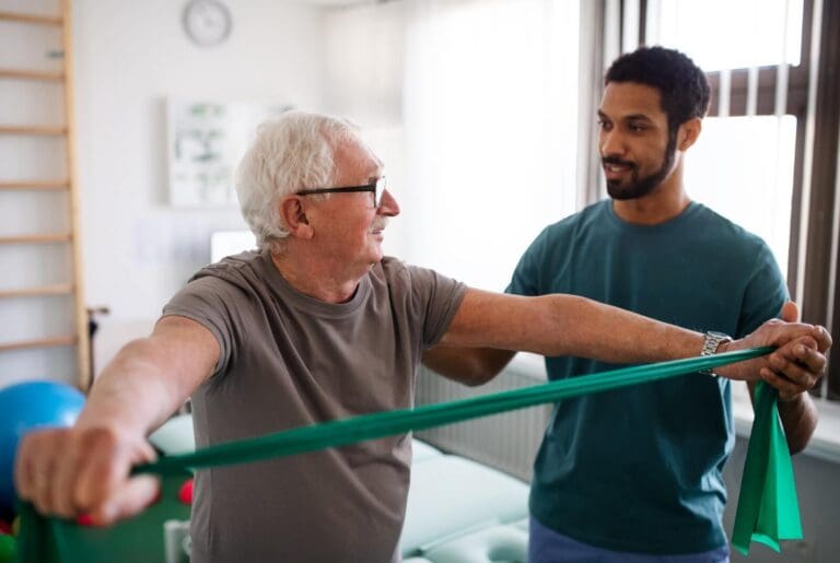 A male physical therapist works with an older adult man. The older man is stretching a resistance band in his hands.