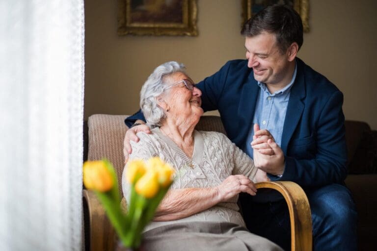 An older adult woman sits in a chair. Her adult son is crouching beside her, holding her hand and placing his arm around her shoulder. They smile at each other.
