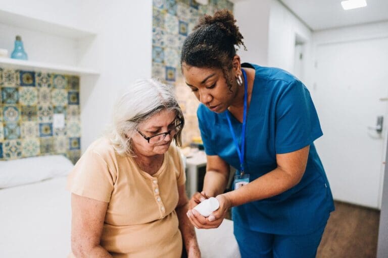 A woman health care worker is standing next to an older adult woman who is sitting on a bed. The health care worker is holding a bottle of medication, showing it to the older woman.