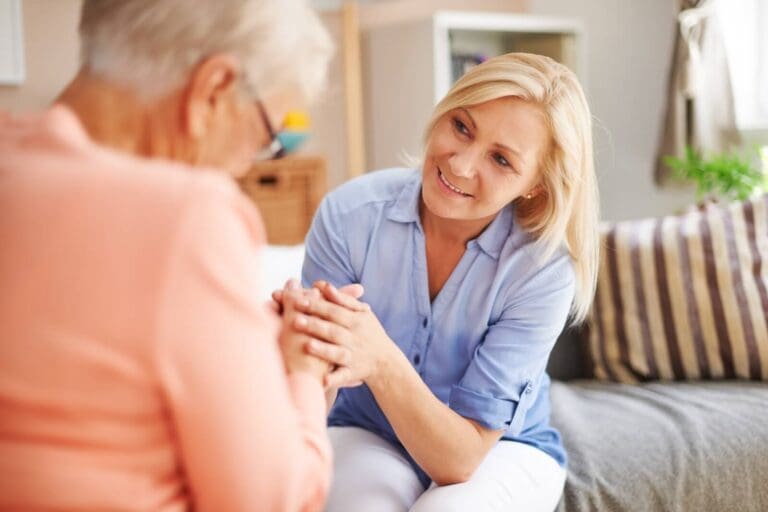 A woman is sitting on a couch with her older adult mother, holding her hands.
