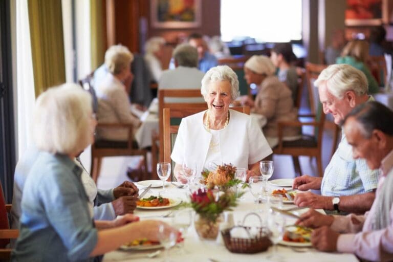 A group of five older adult men and women are sitting at a table in a dining room with food and drinks in front of them. They are happy. There are other table with other older adults in the background.
