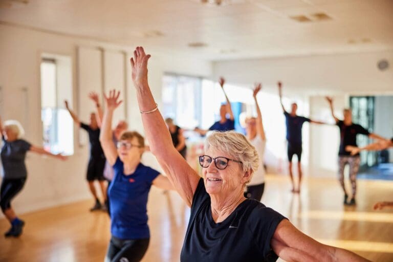 A group of ten or more older adults are standing in a room with no furniture with their arms raised. They are participating in an exercise class.