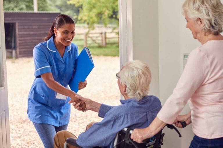 A female home health care provider is walking in the front door of a home. She is shaking the hand of an older adult man who is seated in a wheelchair. An older adult woman stands behind him with her hands on the wheelchair's handles.