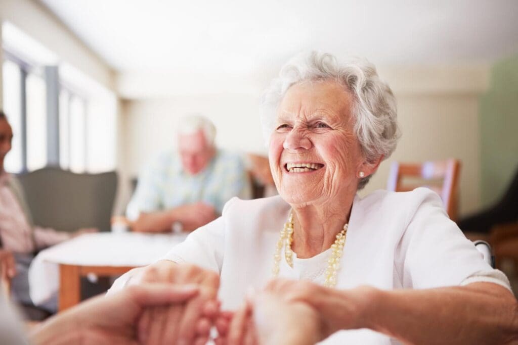 A smiling older adult woman holds the hands of a person who is out of frame. There are two people sitting at a table in the background.