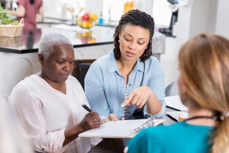 A woman and her older adult mother are sitting at a desk with a woman caregiver.