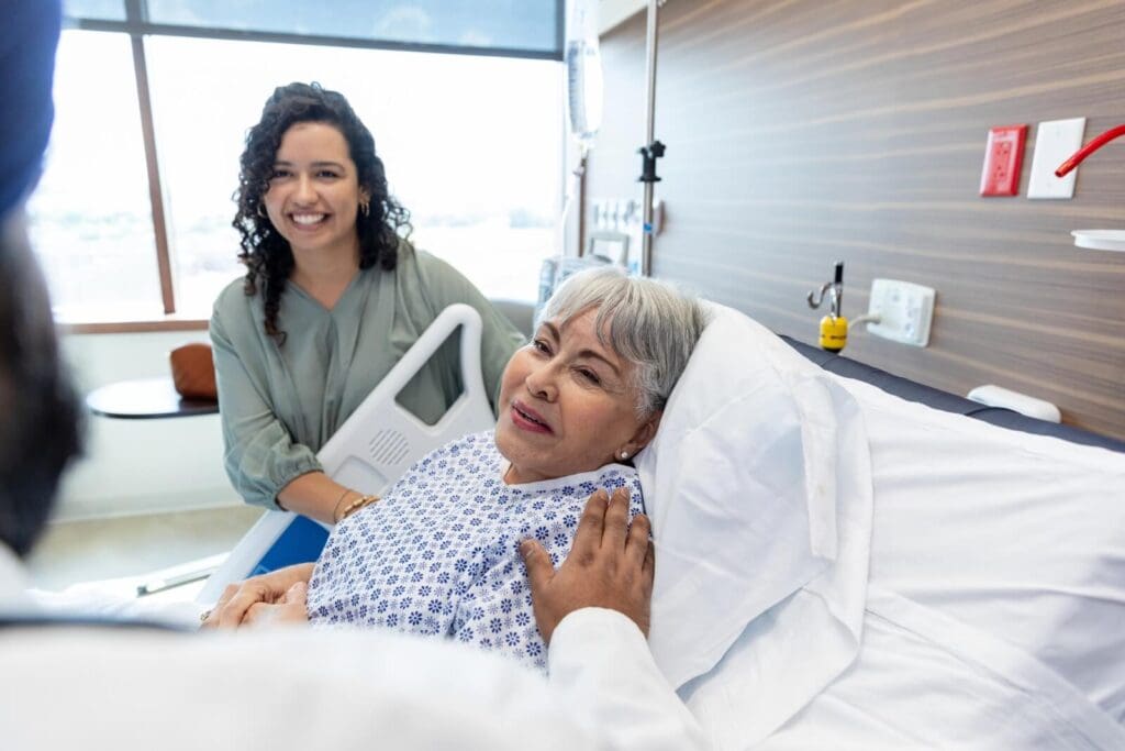 An older adult woman is lying in a hospital bed wearing a hospital gown. Her daughter is standing next to her. They are speaking to a person who is in the foreground.