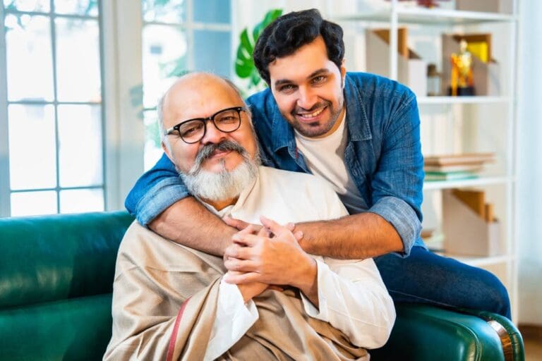 An older adult man sits on a couch. His son is sitting on the arm of the couch next to him with his arms around his father.