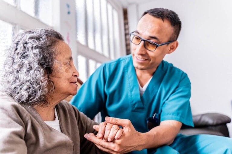 An older adult woman sits next to a male caregiver on a couch. He is holding her hand.