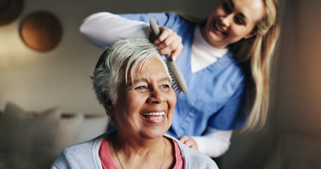 A woman caregiver brushes an older adult woman's hair.