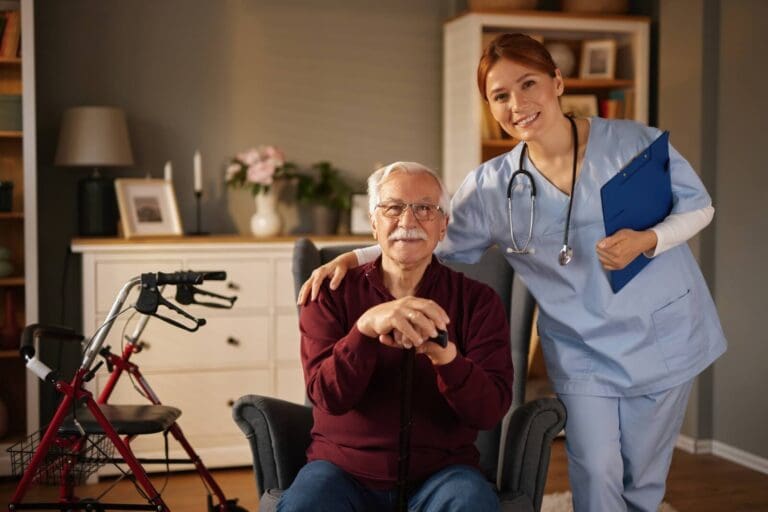 An older adult man sits in a chair holding a cane. A woman health care worker holding a clipboard and wearing a stethoscope is standing next to him with her arm around his shoulders.