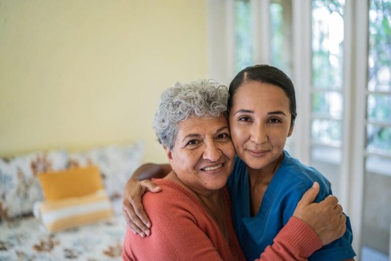 An older adult woman and a woman aide have their arms around each other and are smiling at the camera.