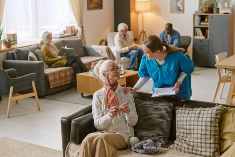 An older adult woman sits on a couch in senior living, working on some knitting. An aide stands behind her with her hand on her shoulder. There are three other older adults on couches and chairs in the background.