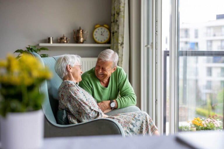 An older adult woman sits in an armchair facing a large window. An older adult man is crouched next to her, smiling at her.