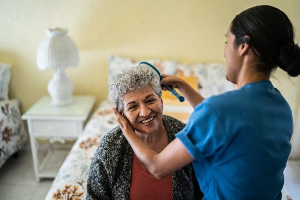 A woman aide brushes the hair of an older adult woman who is seated on a bed.