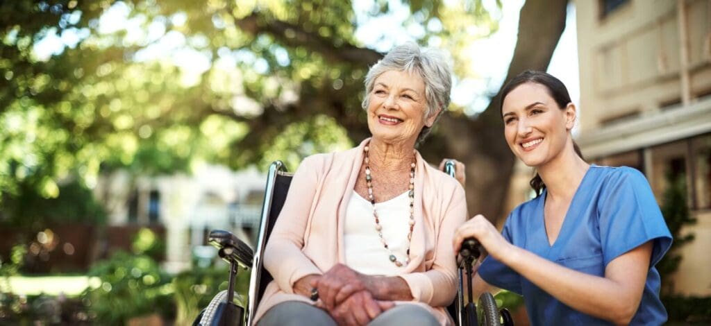 A woman aide crouches next to an older adult woman who is seated in a wheelchair. They are outside.
